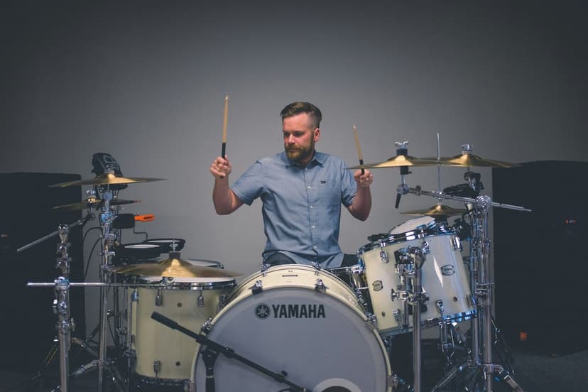 Bearded man in a blue shirt plays a white Yamaha drum set with wooden sticks against a gray studio background.