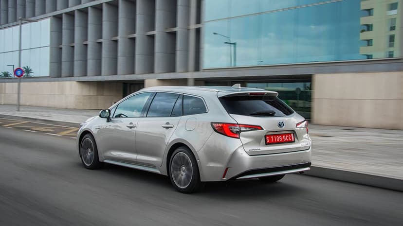 Silver Toyota Corolla wagon driving on a city street in front of a modern glass and concrete building.