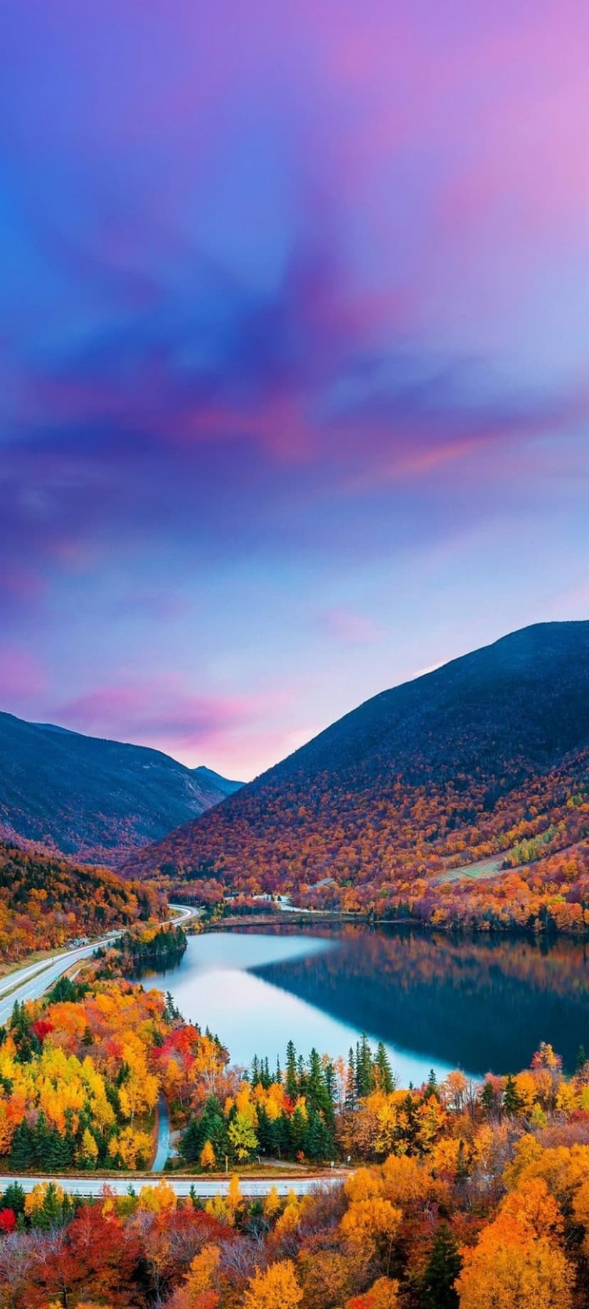 Winding asphalt road through orange autumn forests beside a mountain lake under a vivid purple sunset sky.
