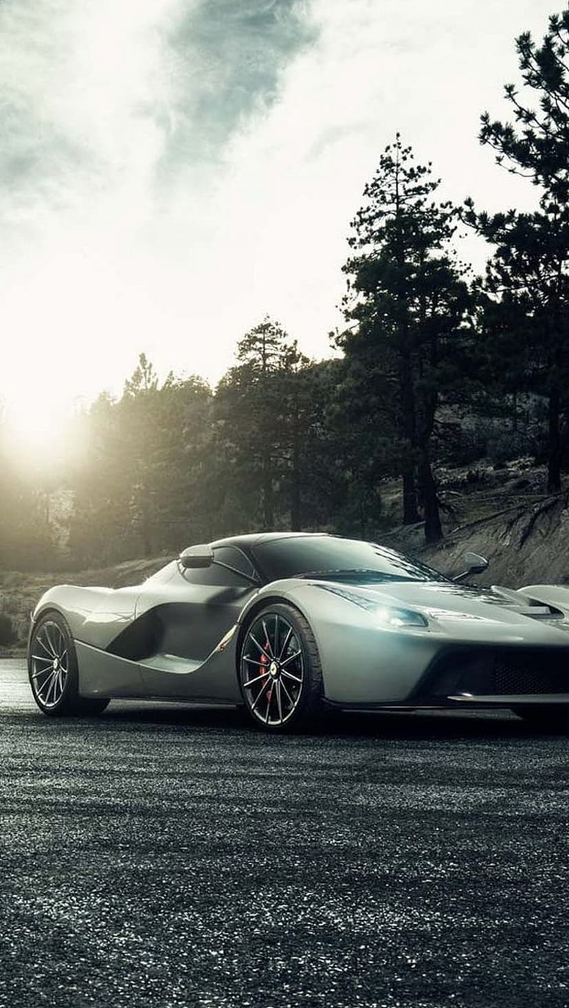 Silver Ferrari LaFerrari on a wet road at sunset with lush green trees and rocky cliffs in the background.