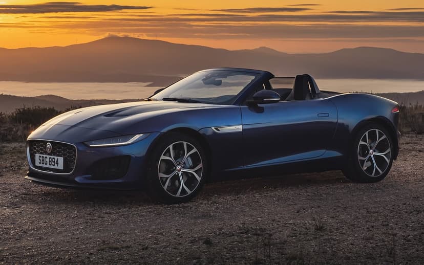 Blue Jaguar convertible parked on a gravel hill at sunset with misty mountains in the background.