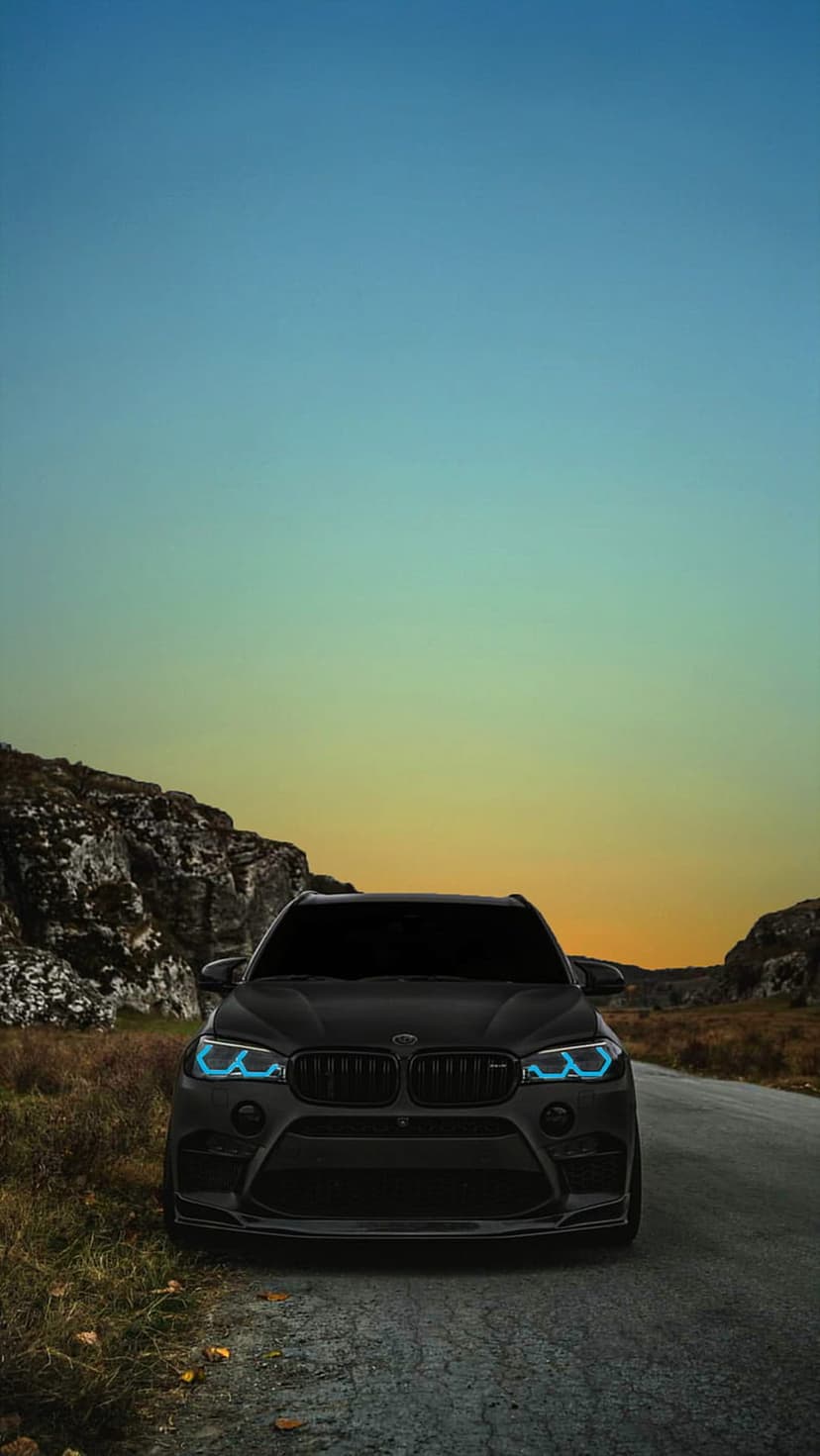 Black BMW X5 M SUV parked on a dirt path among rocks under a golden sunset with glowing blue headlights.