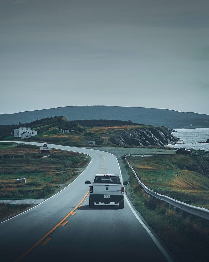 White pickup truck drives along a winding seaside highway past rocky cliffs and misty ocean views under grey skies.