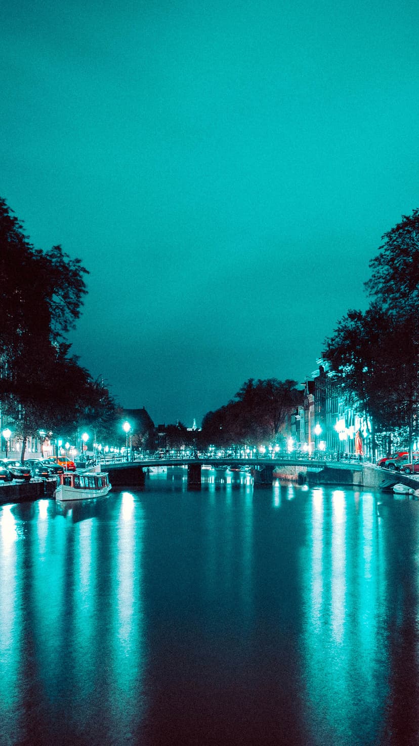 Amsterdam canal bridge illuminated by teal lights reflecting in dark water with docked boats and bare winter trees.