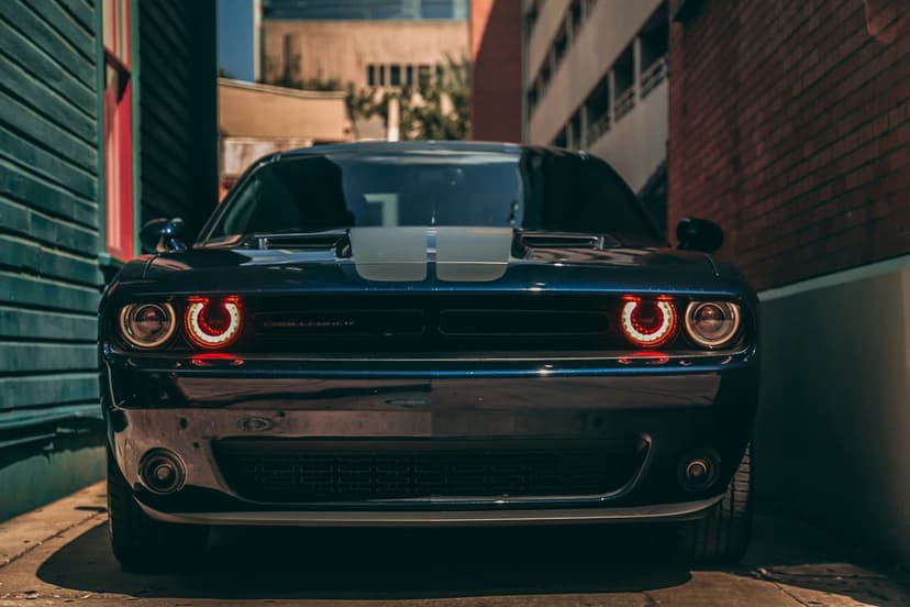 Dark blue Dodge Challenger with glowing red halo headlights and silver racing stripes in a sunlit urban alley.