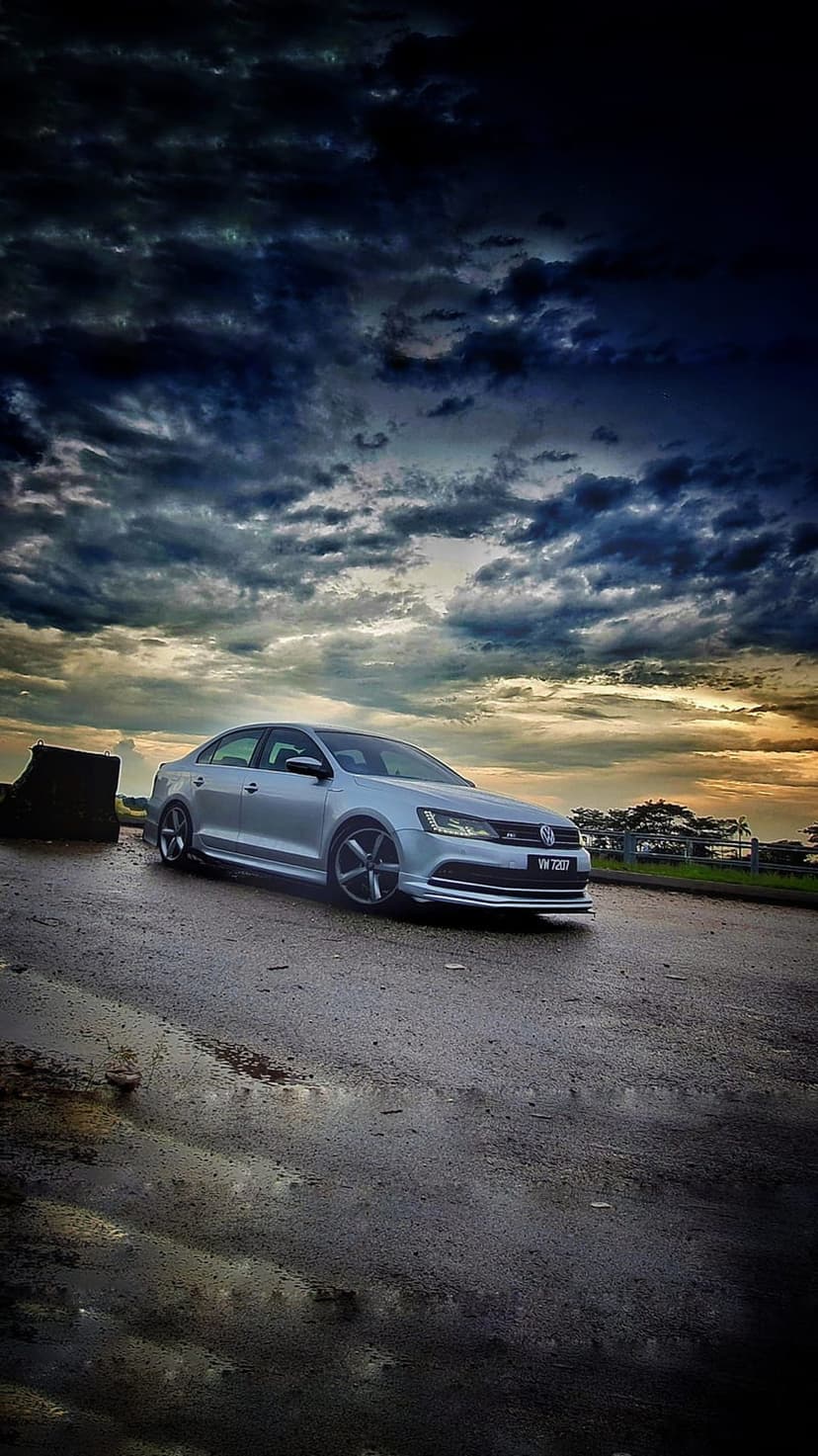 Silver Volkswagen Jetta sedan parked on wet asphalt under dark, dramatic storm clouds with a faint sunset glow.