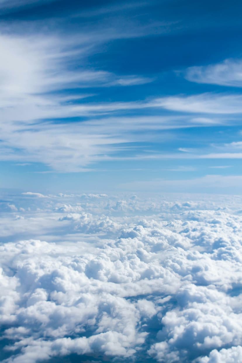 Billowy white cumulus clouds stretch across the atmosphere beneath a vibrant blue sky seen from an aerial view.