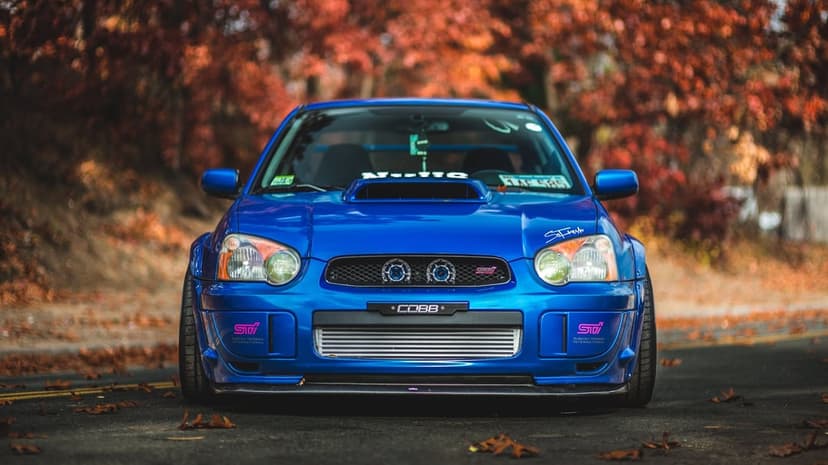 Blue Subaru WRX STi parked on a dark asphalt road with vibrant orange and red autumn trees in the blurred background.