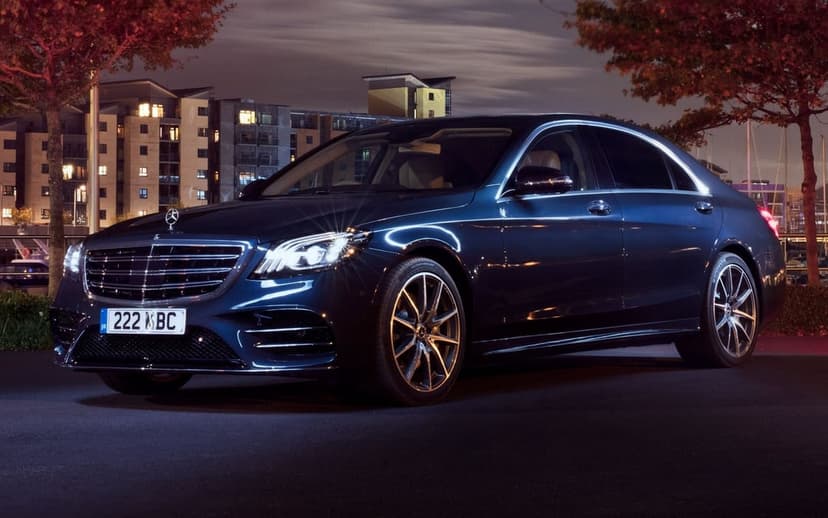 Dark blue Mercedes-Benz S-Class sedan with glowing headlights parked on a city street at night under warm lights.