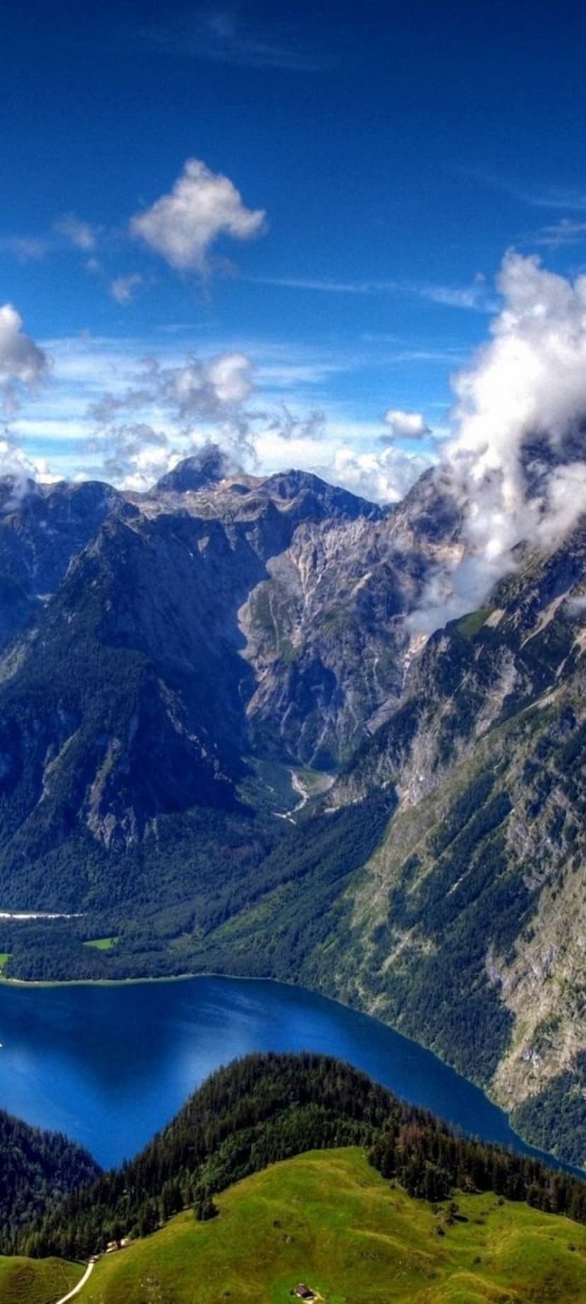 Snow-capped mountains tower above a vibrant blue glacial lake and green forests under a cloudy summer sky.