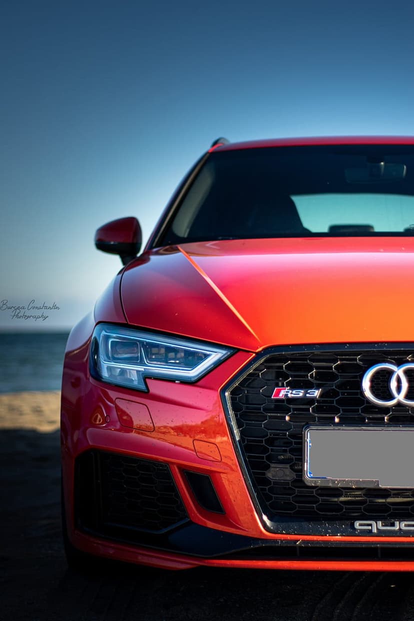 Red Audi RS3 parked on beach sand with ocean waves and a bright blue sky backdrop in a vertical car portrait.