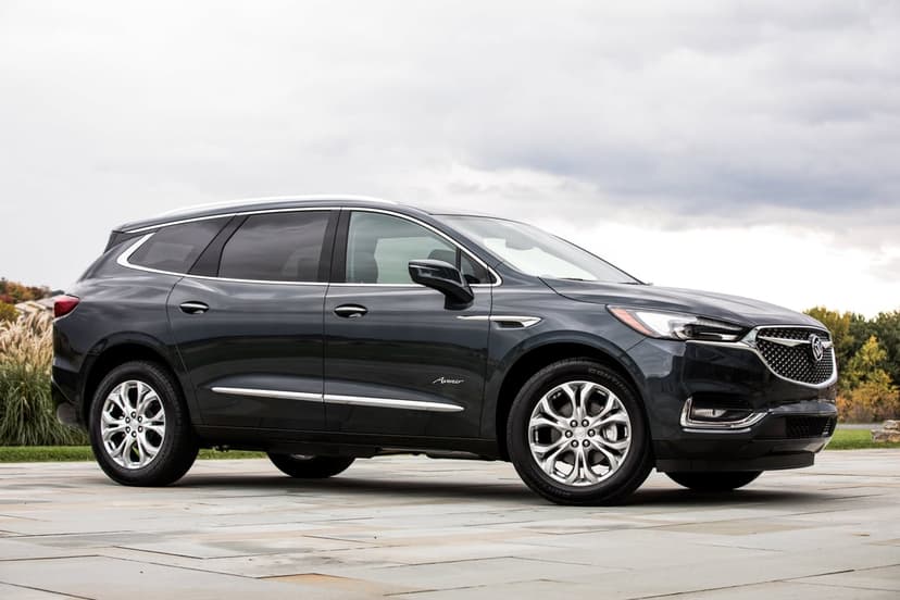 Side view of a dark gray Buick Enclave Avenir SUV on light stone pavers under an overcast sky with distant trees.