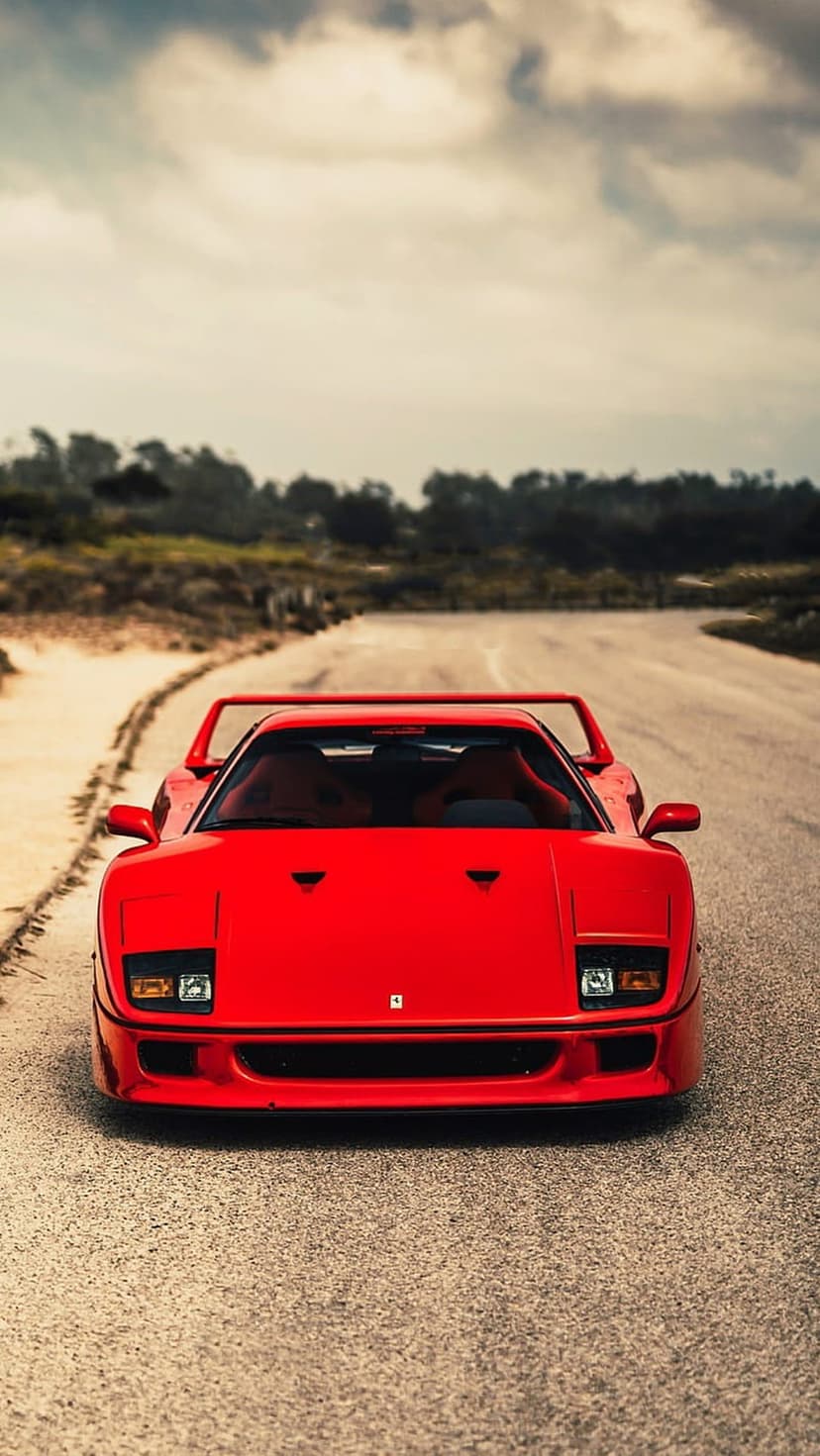 Red Ferrari F40 supercar parked on a dusty dirt road with a large rear spoiler under a gray cloudy sky.