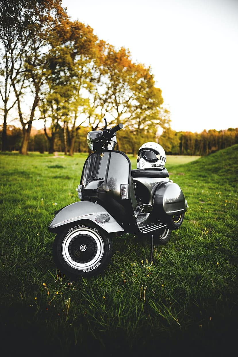 Classic dark scooter with a white helmet and goggles parked in a meadow with yellow wildflowers and trees.