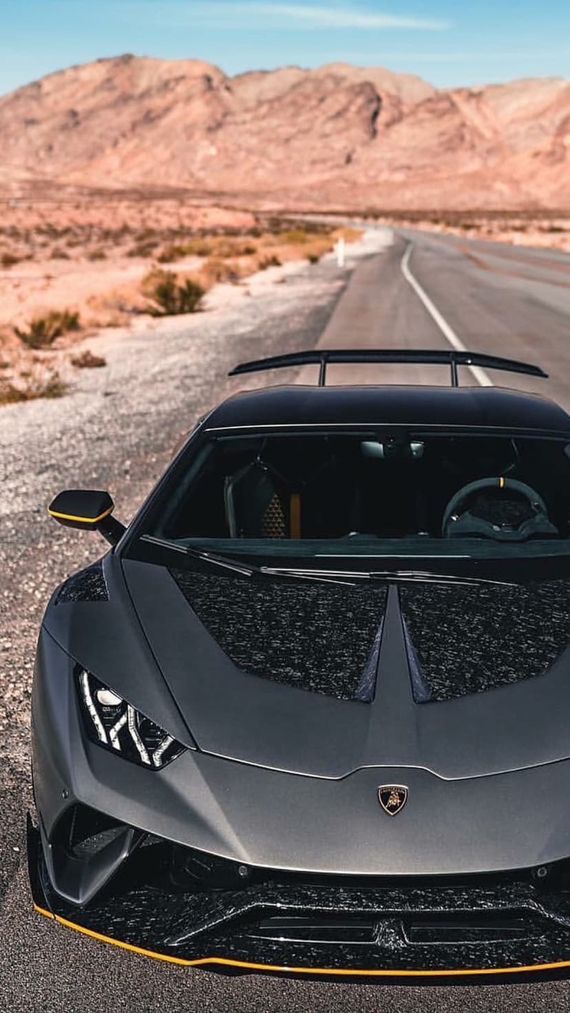 Gray Lamborghini supercar with a rear spoiler parked on an asphalt road surrounded by arid desert mountains.