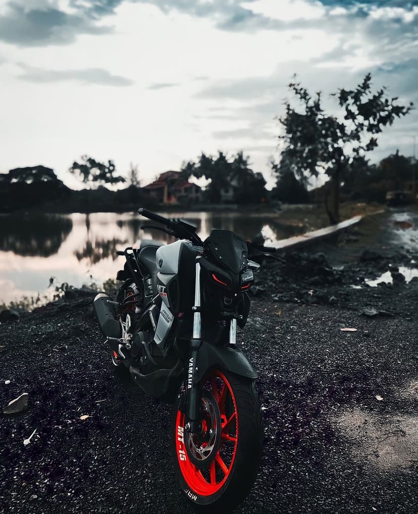Yamaha MT-15 motorcycle with red wheels parked on a rocky beach near a calm lake under a dramatic cloudy sky.