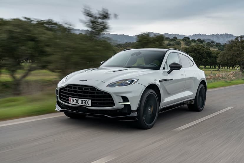 Light grey Aston Martin DBX SUV driving on a paved road with blurred green trees and distant mountains.