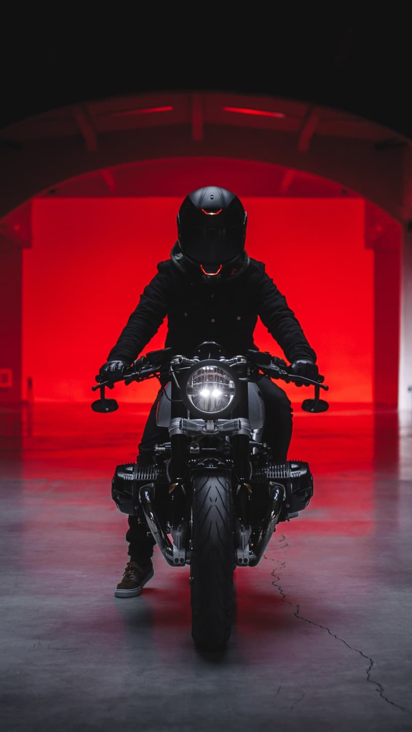Biker in black gear sits on a motorcycle with a round headlight under intense red lighting on a concrete floor.