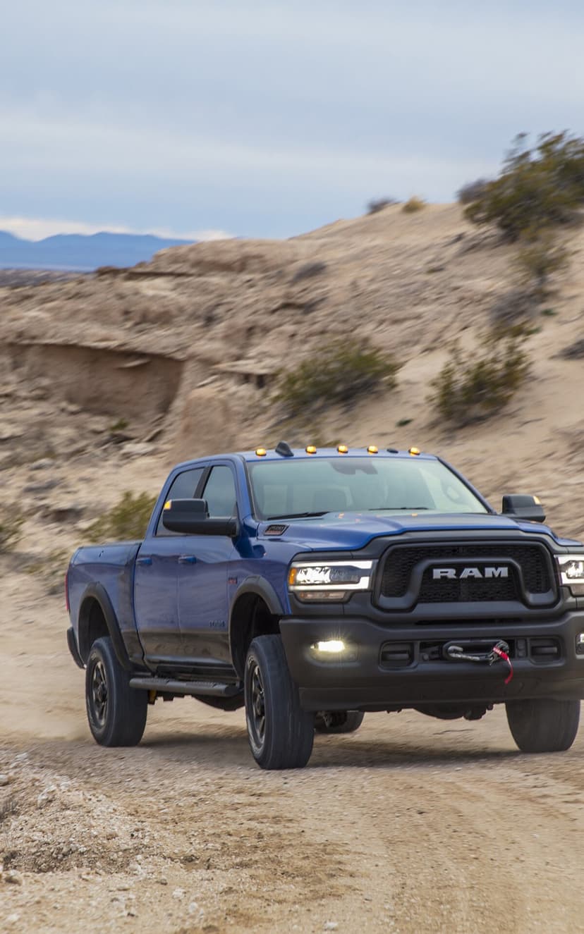 Blue RAM Power Wagon truck driving on a sandy desert path with dust trails and distant dunes.