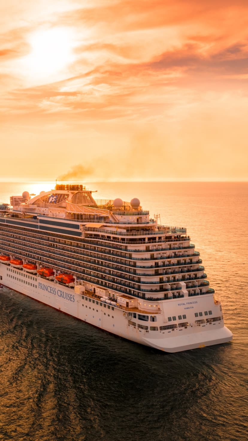 White cruise ship sails across a calm ocean under a bright orange sunset sky with a distant horizon line.