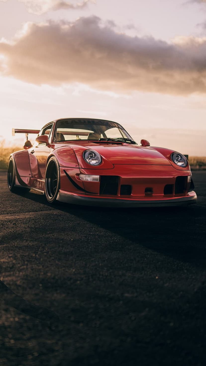 Red Porsche 911 GT3 RS with a widebody kit parked on asphalt during a dramatic sunset with orange and grey clouds.