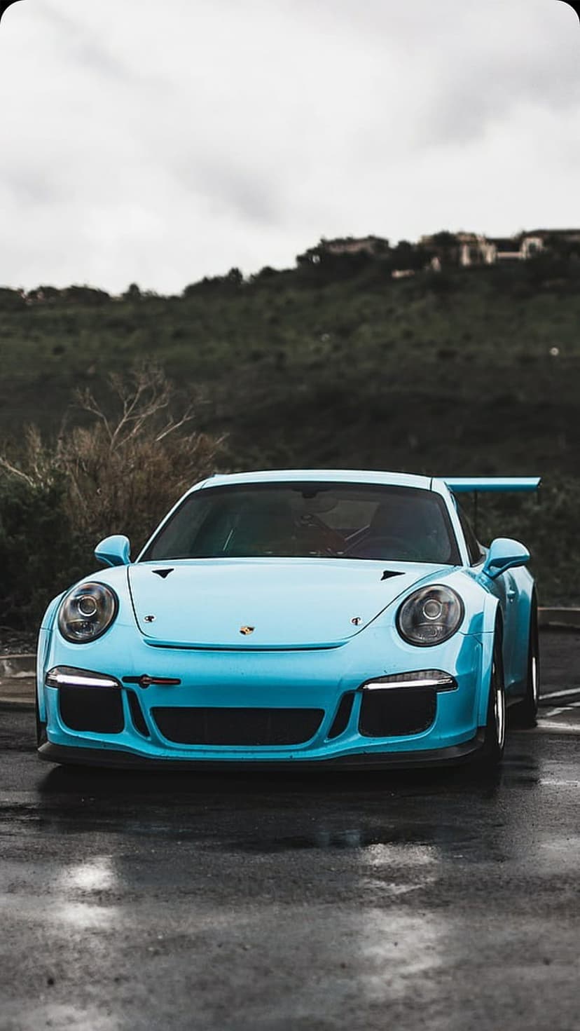 Teal Porsche 911 GT3 RS on damp pavement showing a large rear wing against a blurred misty mountain backdrop.