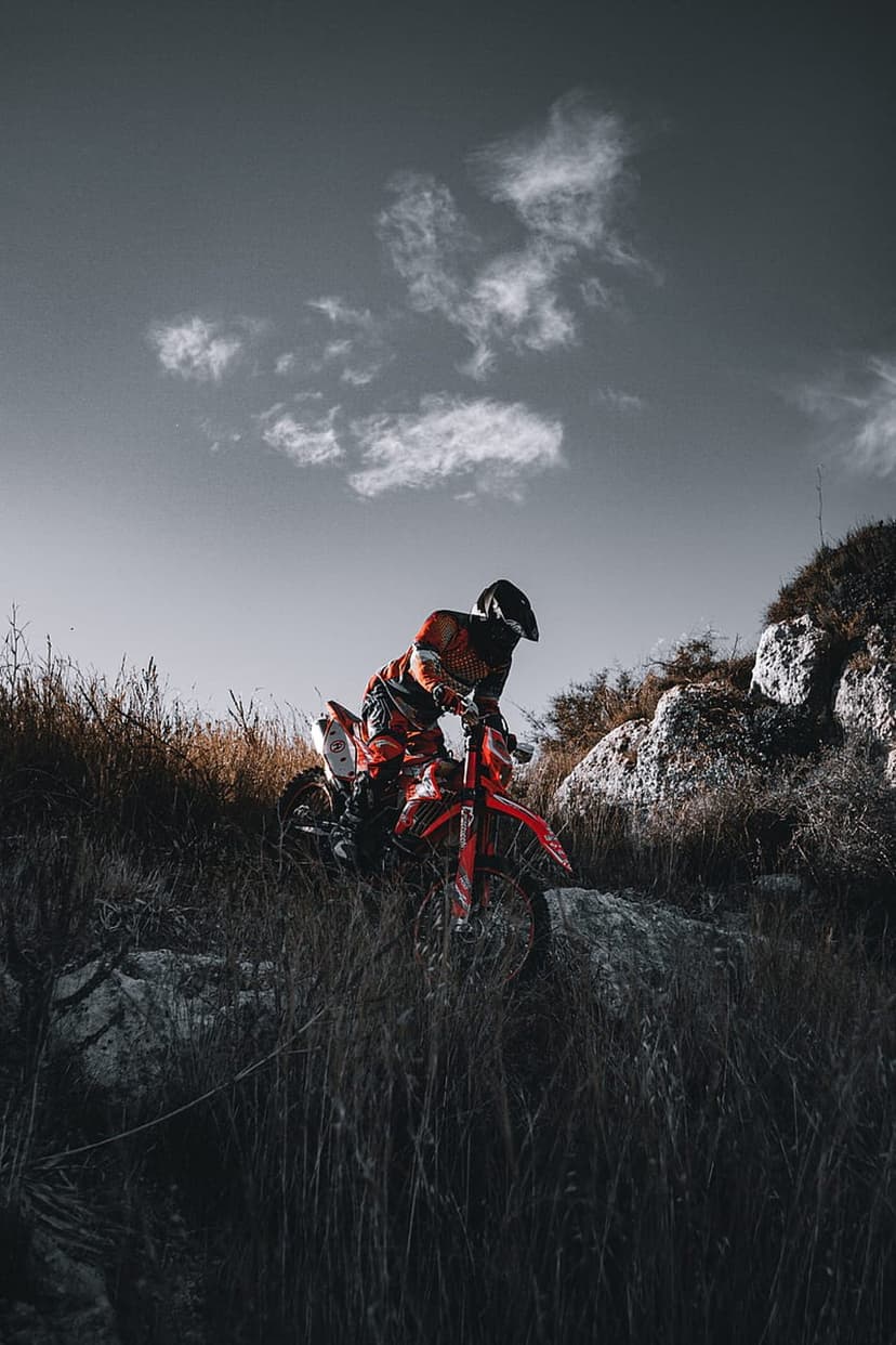 Red motocross bike rider navigates sharp grey boulders and dry grass beneath a vast, cloudy, dramatic sky.