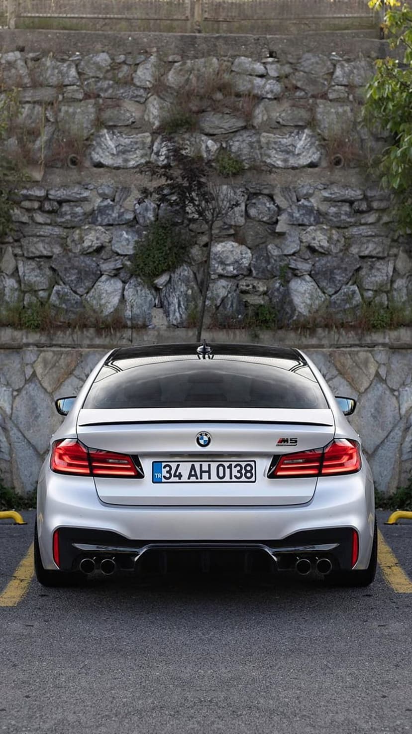 Silver BMW M5 rear view parked by a grey stone wall with visible quad exhausts and red taillights in daylight.