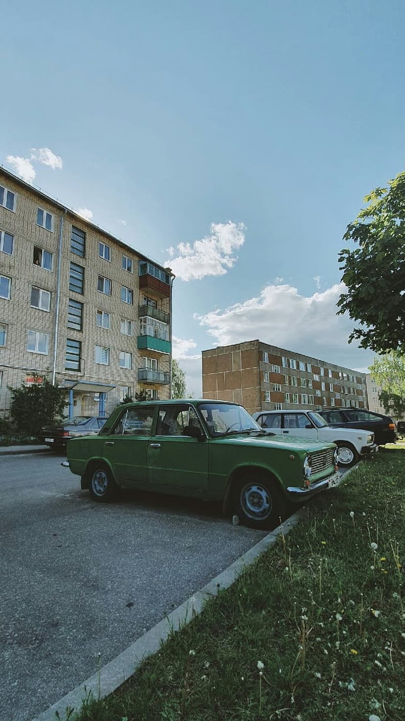 Green vintage car parked on grass before tall white apartment blocks under a clear blue sky with fluffy clouds.