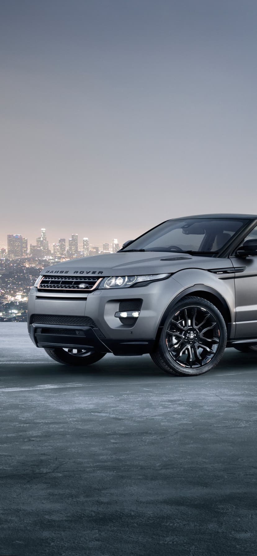 Matte gray Range Rover SUV with black rims on concrete against a distant glowing city skyline at night.