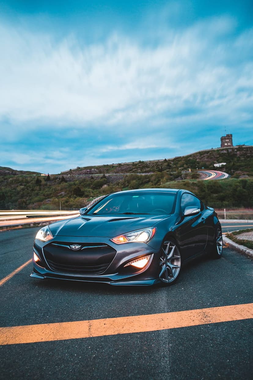 Hyundai Genesis Coupe with lit headlights on a mountain road at dusk near a stone tower and cloudy sky.