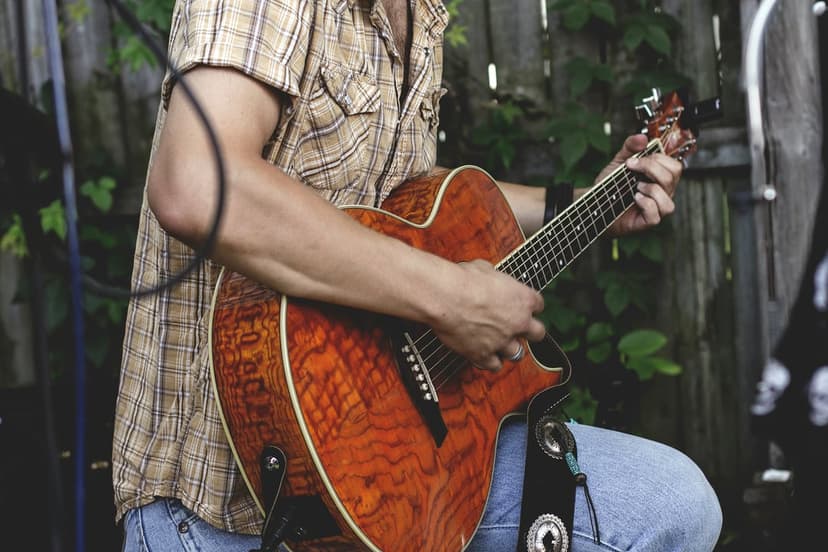 Man in a plaid shirt plays a polished acoustic guitar outdoors with garden greenery and a wooden fence behind him.