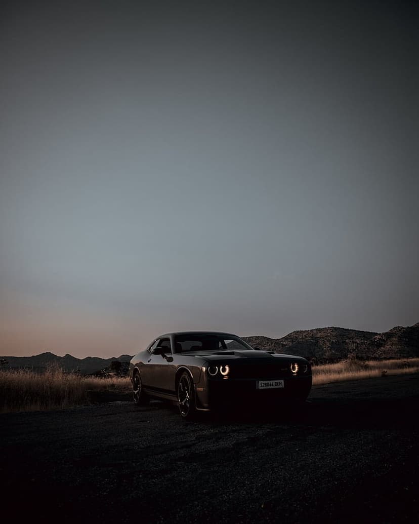 Dodge Challenger with glowing headlights parked on a dirt trail against a sunset mountain range backdrop.