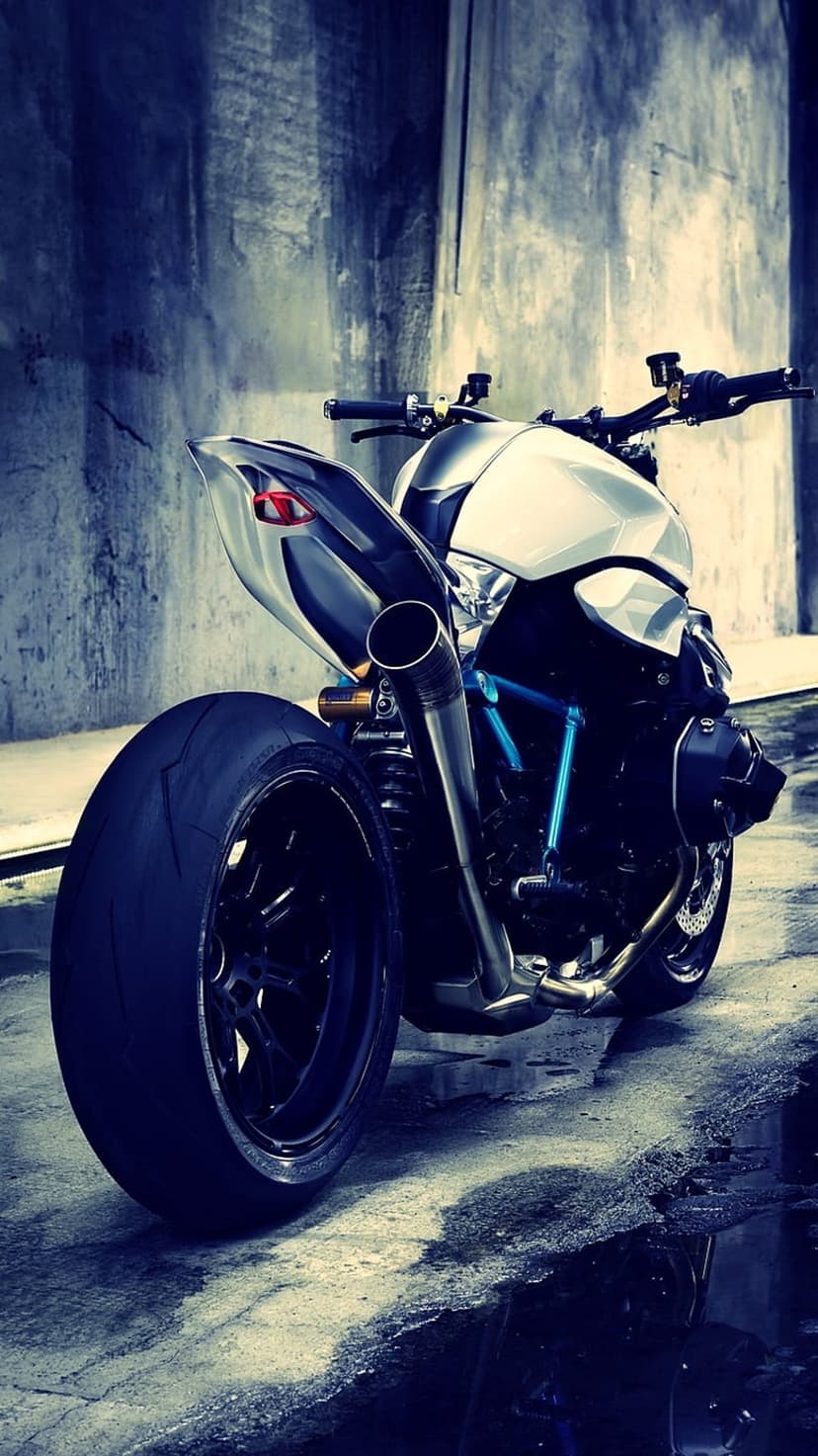 Custom white sportbike with a wide tire and blue accents parked against a rough concrete wall on wet pavement.