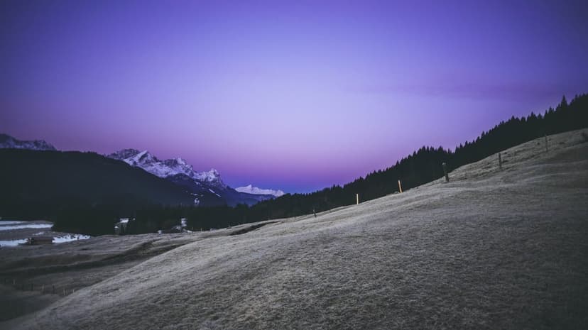 Snow-capped mountains and a grassy hill sit beneath a deep purple twilight sky during a calm alpine evening.