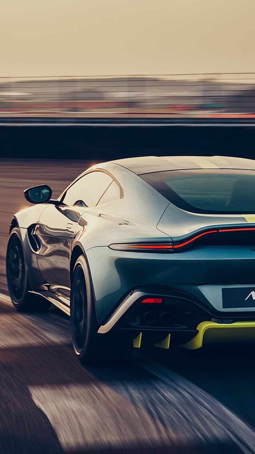 Dark teal Aston Martin Vantage with glowing red taillights speeds on a race track under a hazy sunset sky.