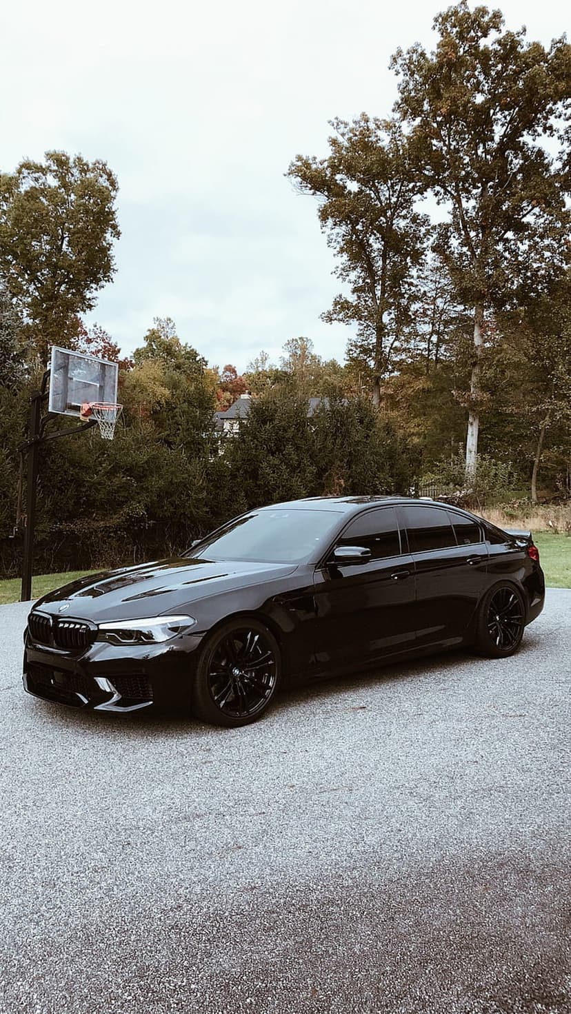 Black BMW M5 sedan with tinted windows and dark rims parked on an asphalt driveway near green trees and a hoop.