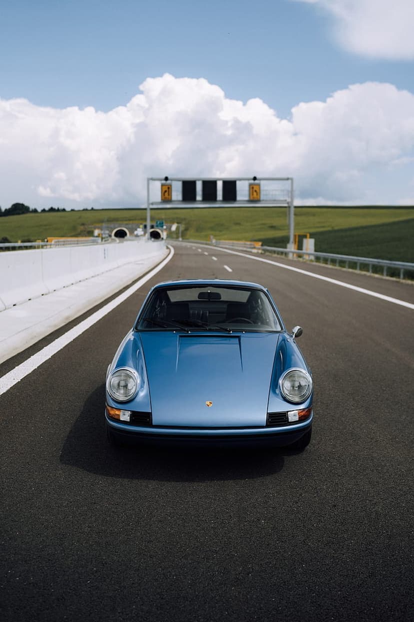 Blue Porsche 911 driving on a highway with green fields and fluffy clouds under a bright summer sky.
