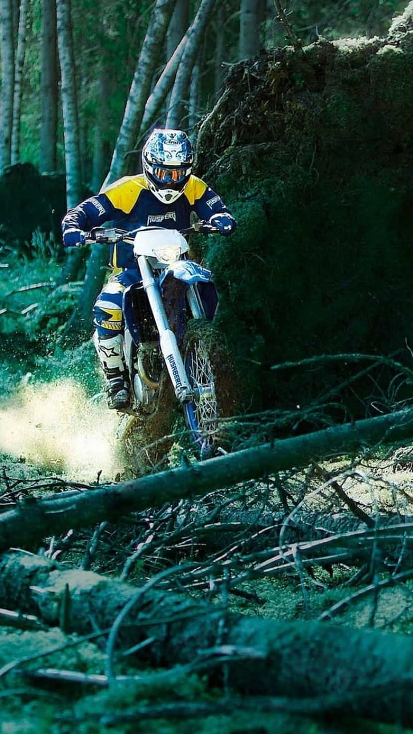 Enduro rider in yellow and blue gear navigates a dirt bike over fallen logs on a dusty, wooded forest trail.