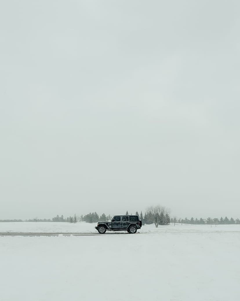 Dark Jeep Wrangler driving across a snow-covered field under a grey overcast sky with a line of bare trees.