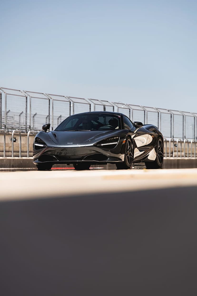 McLaren 720S supercar in dark gray parked on a sunlit racetrack under a clear blue sky with safety fences.