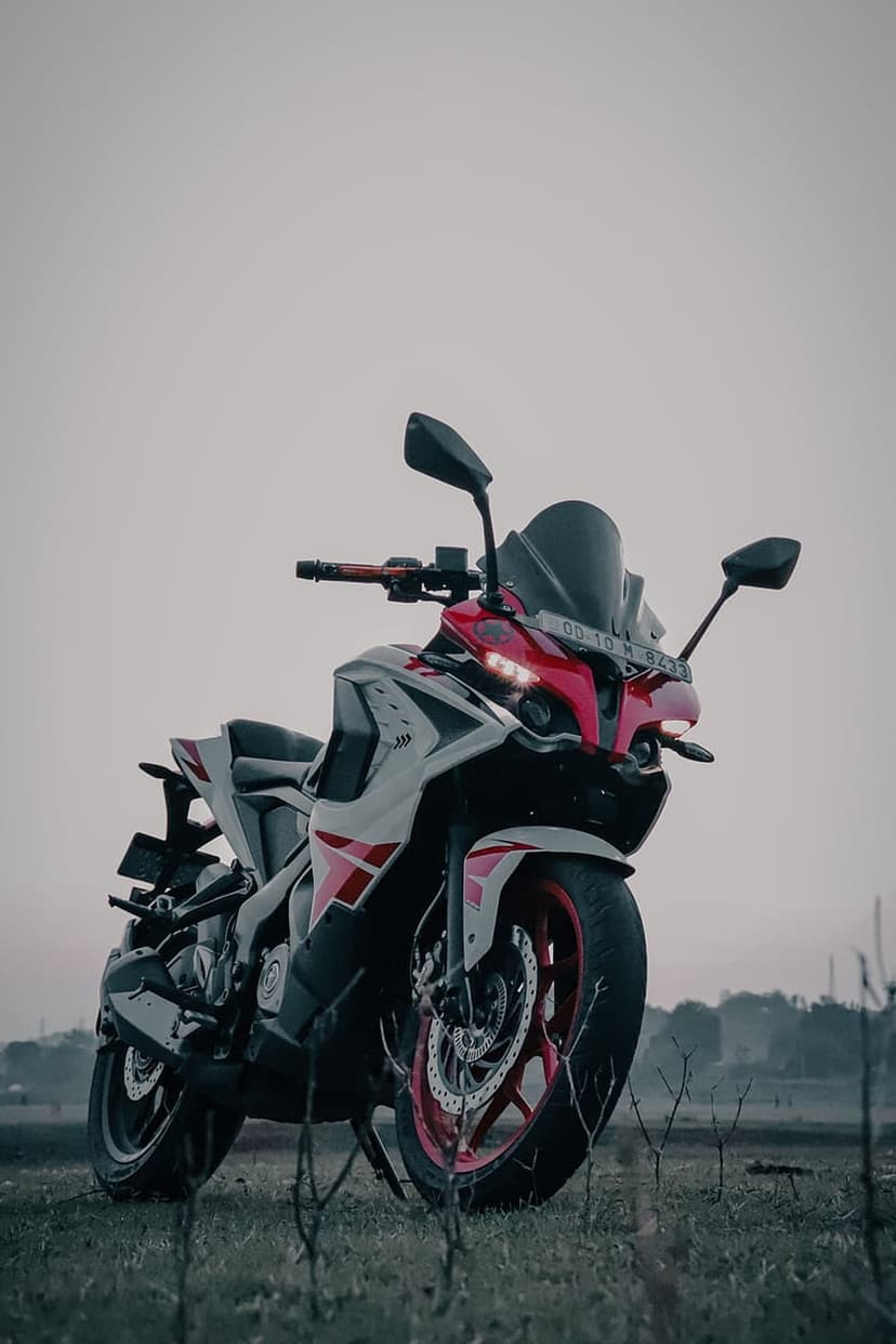 Bajaj Pulsar RS200 motorcycle with red alloy wheels parked in a grassy field under a cloudy, atmospheric sky.