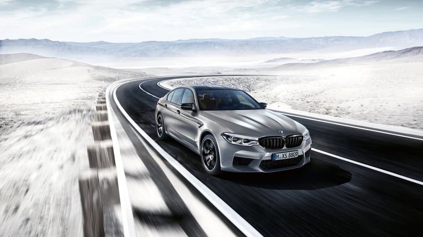Silver BMW M5 F90 driving on a curved asphalt road through a sunlit desert landscape with mountain views.