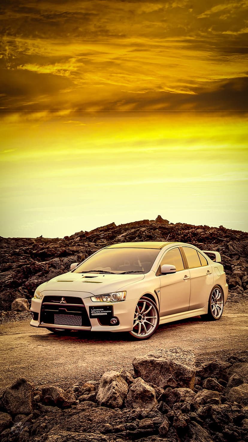 White Mitsubishi Lancer Evolution X sedan on a rocky landscape under a dramatic golden and orange sunset sky.