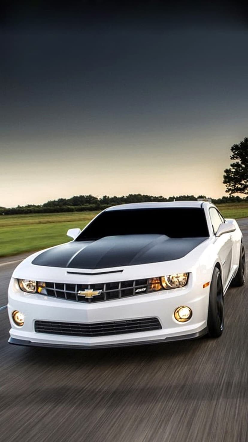 White Chevrolet Camaro with black racing stripes and lit headlights speeds on a road under a moody sunset sky.