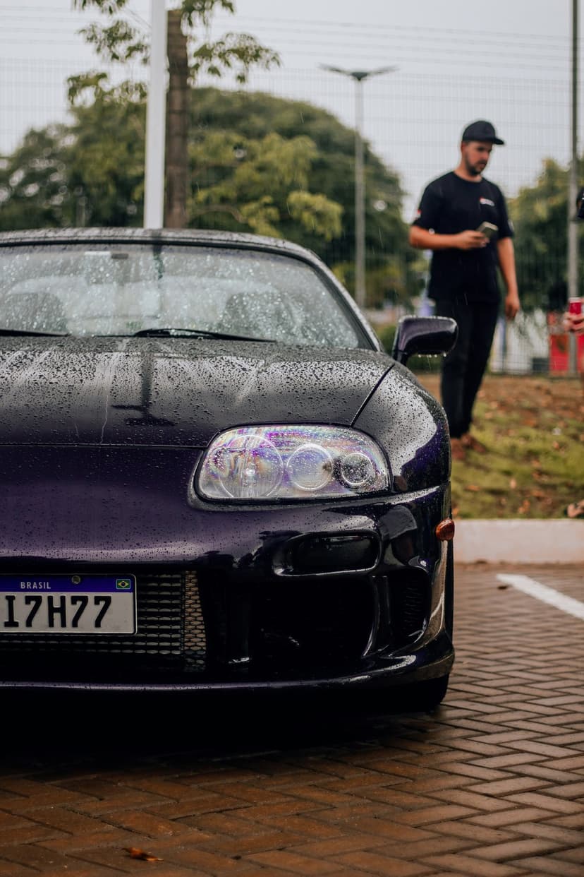 Purple Toyota Supra sports car covered in raindrops parked on wet pavement with a person in the background.