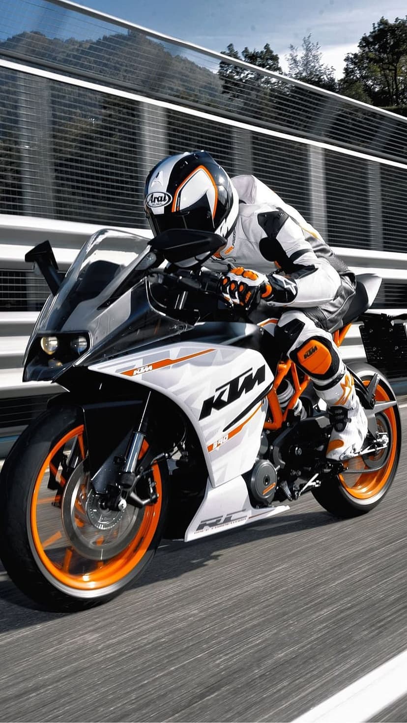 KTM motorcycle rider in white and orange racing gear speeds across a blurred asphalt track under bright sunlight.