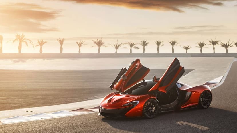 Orange McLaren P1 with open butterfly doors on a racetrack at sunset with palm trees in the background.