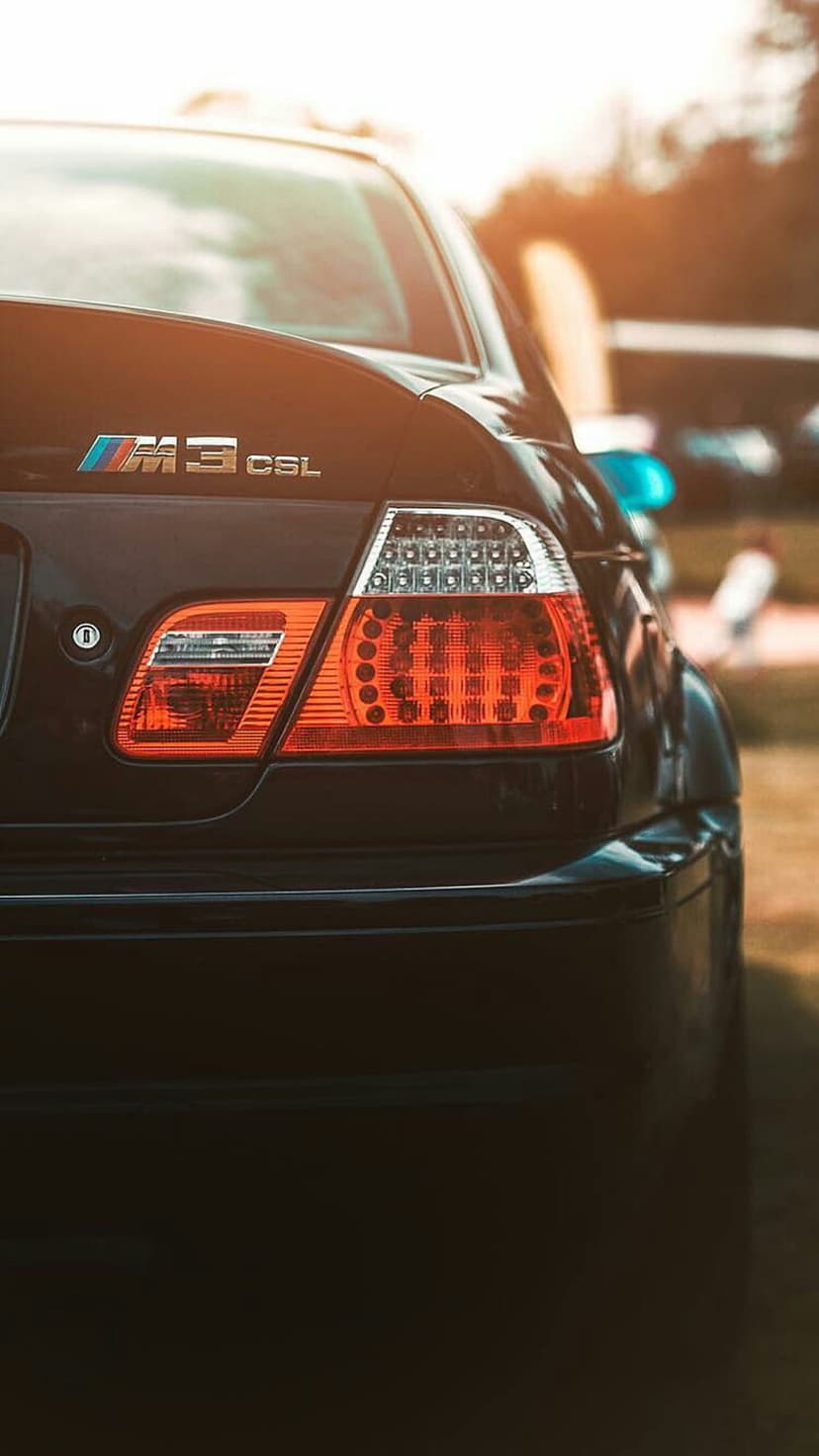 BMW M3 CSL rear tail light and badge illuminated by warm golden hour sunlight on a black metallic car body.