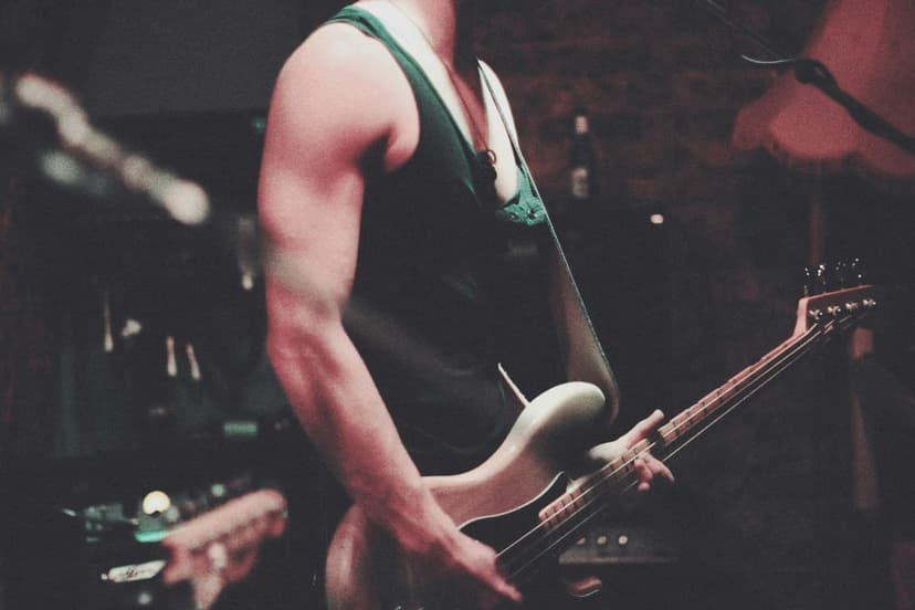 Musician plays a light bass guitar wearing a dark green tank top on a dimly lit stage during a live concert.